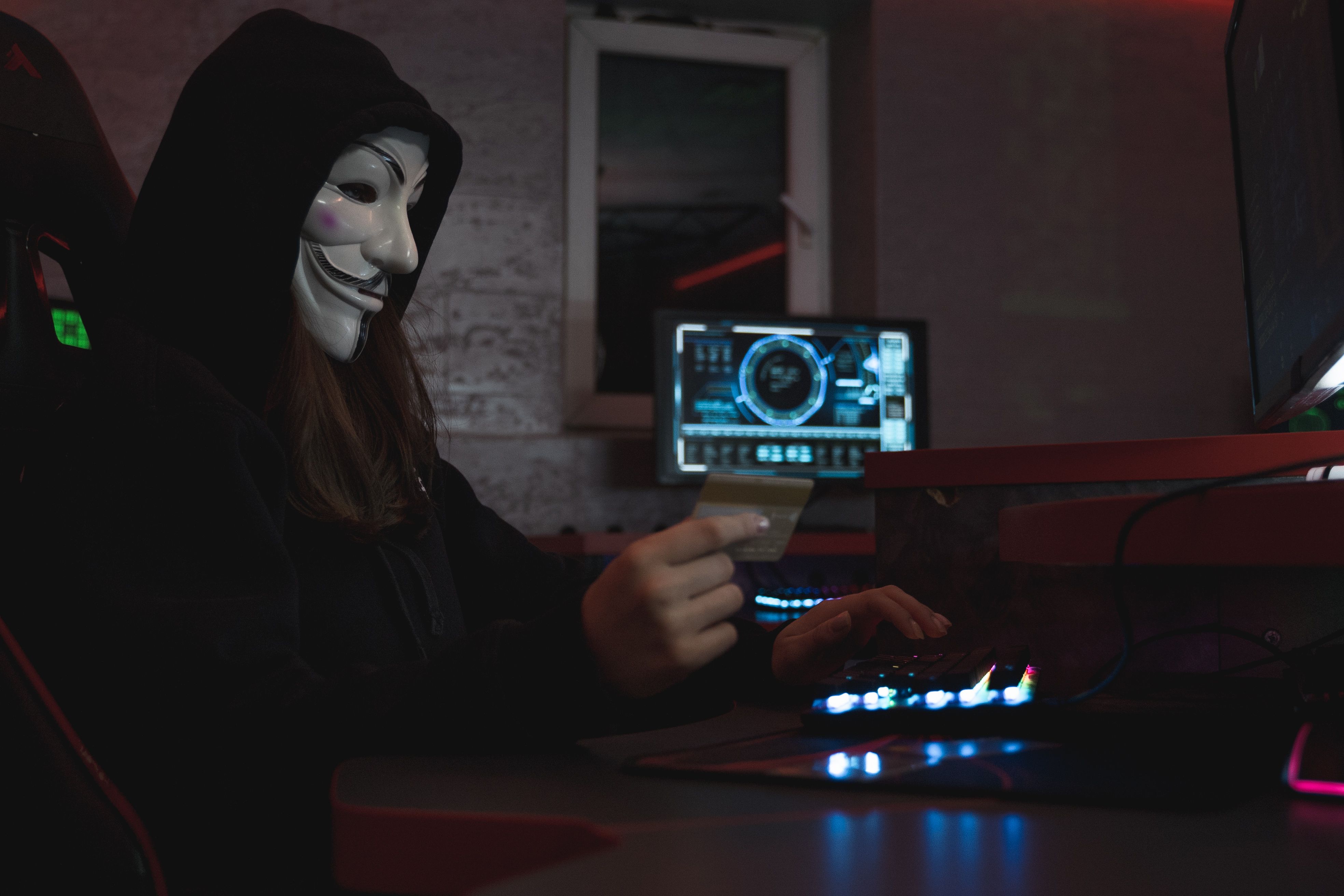 Masked hacker holding a credit card at a computer, representing credit card fraud and identity theft from data breaches